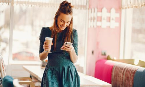 beautiful and stylish woman with long hair and in a green dress standing in a cafe at the table and drinking coffee and using the phone